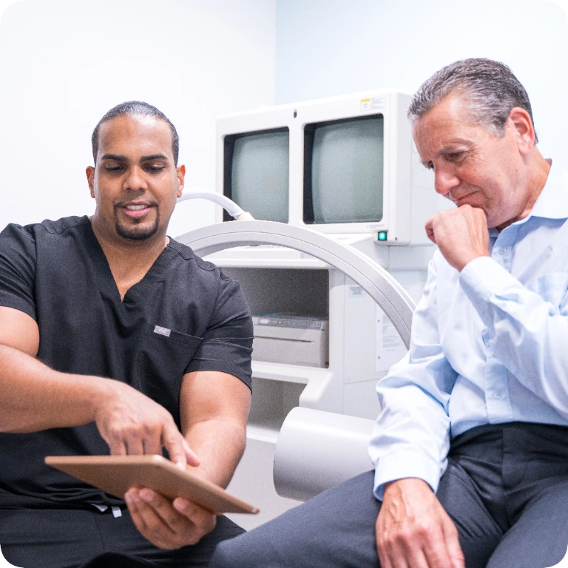 Dr. Jeffrey Deygoo consults with a patient in a clinical setting, reviewing information on a tablet. A C-arm fluoroscopy machine is positioned in the background.