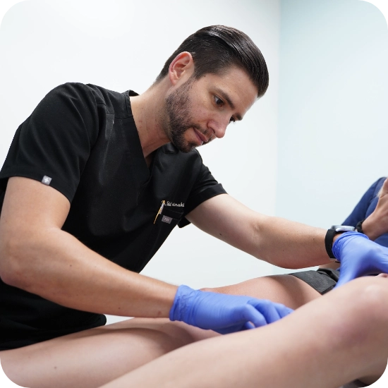 Double certified Dr Kamran Saraf Dr. Todd Kobrinski, MD performs a Duplex Ultrasound examination on a patient's leg to assess for venous insufficiency. The patient wears blue compression therapy wraps on the lower legs during the evaluation. The clinical setting features white walls and medical equipment typical of a vein treatment facility.