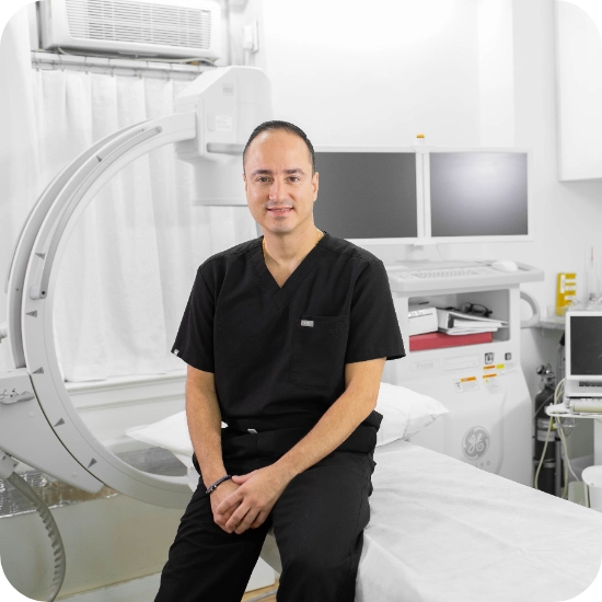 Double certified Dr Kamran Saraf Professional headshot of Dr. Juan Montoya posed with arms crossed against a clinical background with modern machines, wearing a black uniform. This formal studio portrait is suitable for medical practice marketing and professional representation.