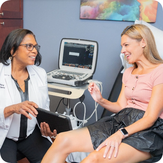 Double certified Dr Kamran Saraf Dr. Lisa Alford is consulting with a patient in a clinical office, using a tablet to review information. The patient is seated on an examination table while Dr. Alford explains the treatment plan.