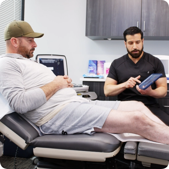 Double certified Dr Kamran Saraf Dr. Andrew Cortes consults with a male patient in a clinical exam room, using a tablet to review a diagnosis or treatment plan. An ultrasound machine is positioned nearby, indicating a venous evaluation is taking place.