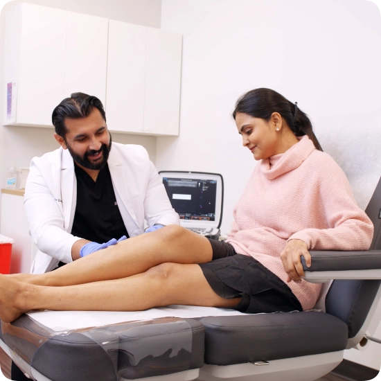 Double certified Dr Kamran Saraf Dr. Sahil Patel, wearing a white coat and blue medical gloves, performs a physical examination on a patient's leg in a clinical setting. The patient is seated on an exam chair while Dr. Sahil Patel assesses the lower extremity, with an ultrasound machine positioned in the background.