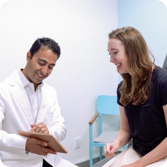 Double certified Dr Kamran Saraf Dr. Kamran Saraf is consulting with a female patient in a clinical examination room. The doctor is showing the patient information on a tablet, and both are smiling during the positive interaction.