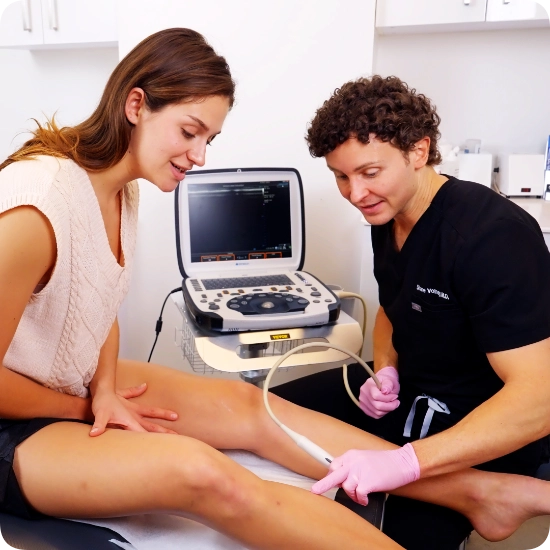 Double certified Dr Kamran Saraf Dr. Shane Volney performs a Duplex Ultrasound examination on a patient's leg to assess for Varicose Veins and Venous Reflux while the patient sits on an examination table. A colleague observes the procedure in this clinical VTC setting. The Ultrasound Machine is positioned nearby to visualize the venous structures and blood flow patterns.