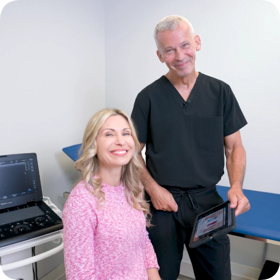 Double certified Dr Kamran Saraf Dr. Martin Tyson stands with a patient in a clinical examination room, both smiling at the camera. He holds a tablet displaying medical imagery, with an ultrasound machine positioned nearby.