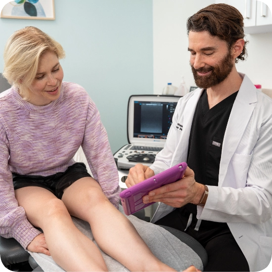 Double certified Dr Kamran Saraf Dr. Shaun Cole consults with a female patient in a clinical examination room, reviewing findings using a handheld device. A Duplex Ultrasound machine is visible in the background, indicating a comprehensive vascular consultation for venous assessment and treatment planning.