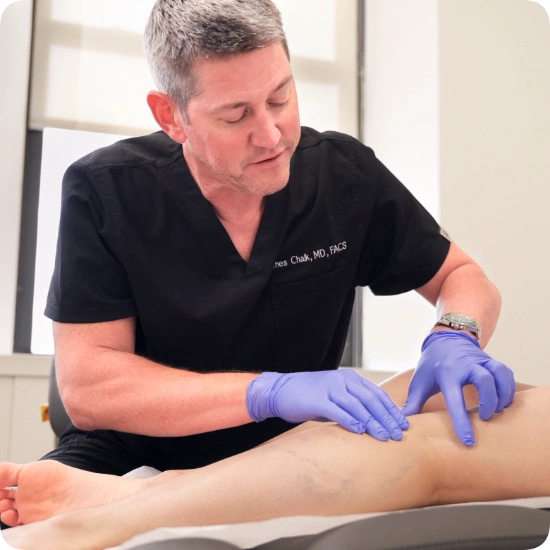 Double certified Dr Kamran Saraf Dr. James Chalk is performing a clinical examination on a patient's leg in a medical office setting. Wearing blue medical gloves, he is carefully assessing visible superficial veins on the patient's lower extremity.