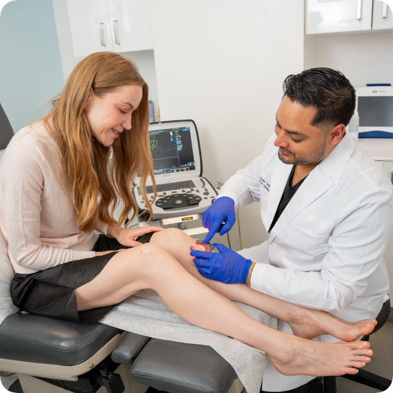 Double certified Dr Kamran Saraf Dr. Ryan Sadloo is performing a medical examination on a female patient's leg in a clinical setting. He is using a Veinlite to illuminate the veins on her lower leg, with an ultrasound machine positioned in the background.