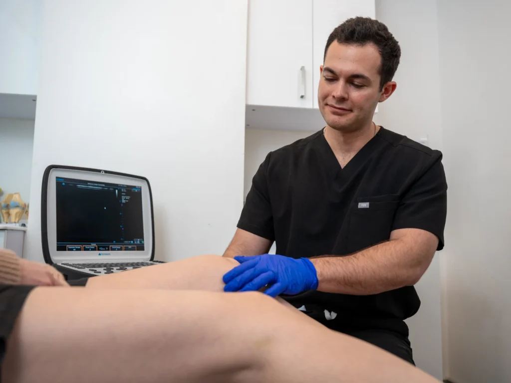 Dr. Kyle Mele is performing a clinical examination on a patient's leg. He is using an ultrasound machine in the background to assist with the diagnostic imaging of the lower extremity.