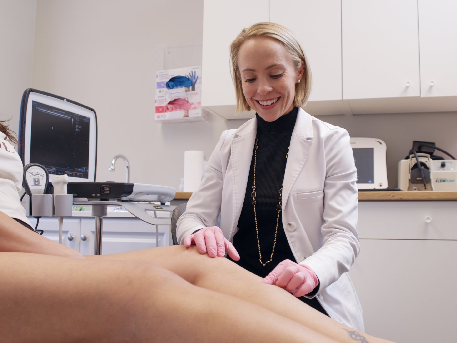 Dr. Laura Lombardi, wearing pink medical gloves, performs a physical examination on a patient's leg in a clinical setting. An ultrasound machine is positioned in the background, facilitating the vascular consultation.