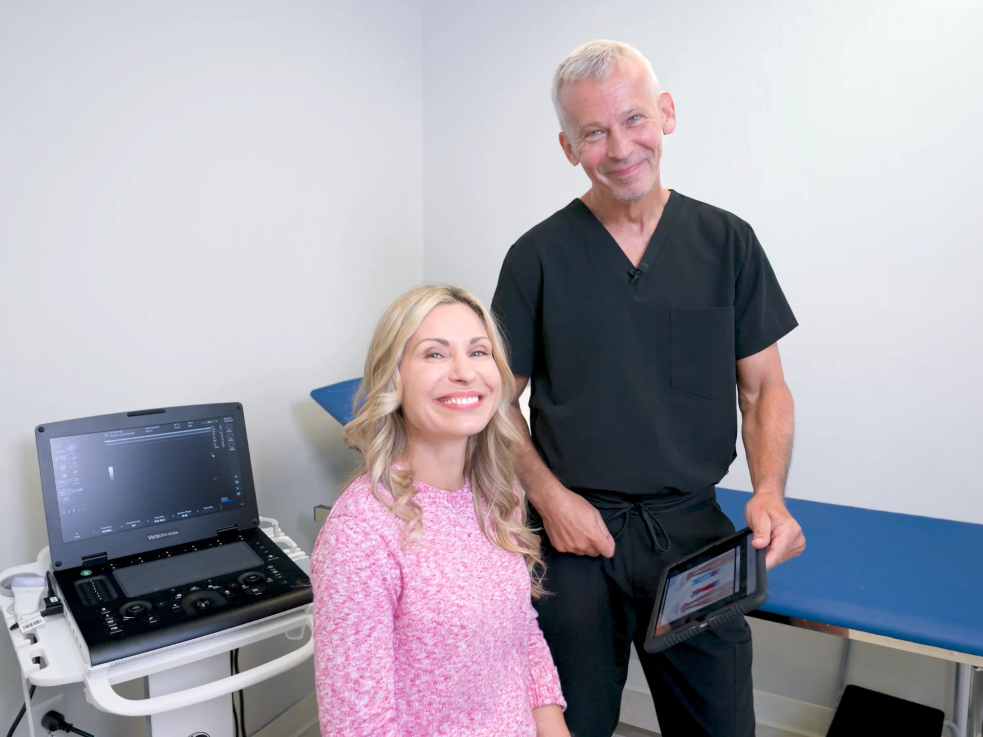 Dr. Martin Tyson stands with a patient in a clinical examination room, both smiling at the camera. He holds a tablet displaying medical imagery, with an ultrasound machine positioned nearby.