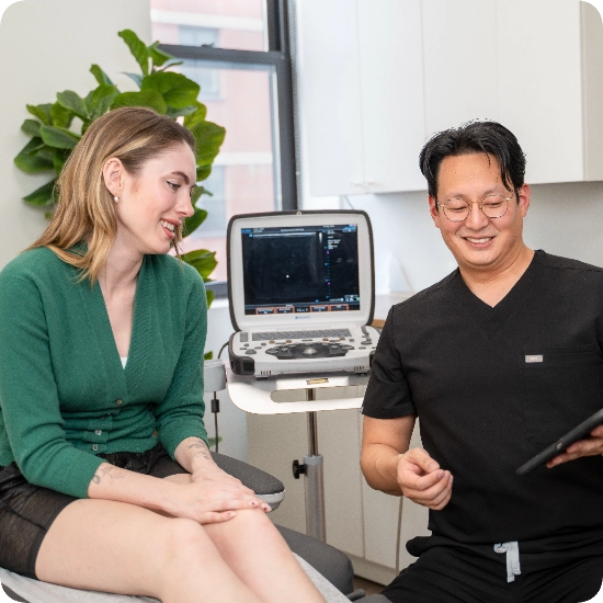 Double certified Dr Kamran Saraf Dr. Benjamin Kum is consulting with a female patient in a clinical office setting, reviewing diagnostic information on a tablet. An ultrasound machine is positioned in the background, highlighting the focus on diagnostic imaging.