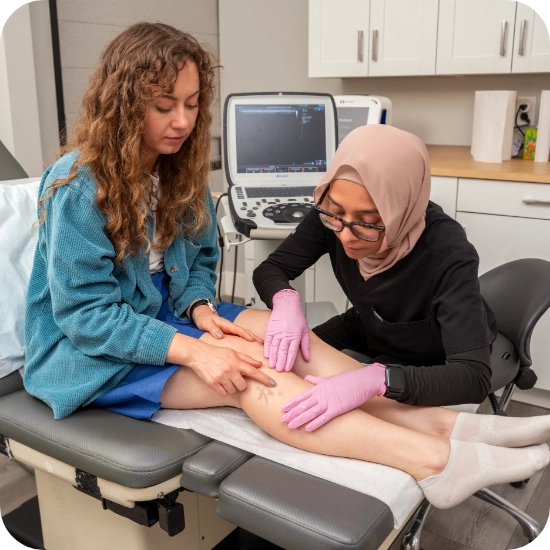 Double certified Dr Kamran Saraf Dr. Anee Khan performs a physical examination of a patient's lower leg and calf area for venous assessment while the patient sits on an examination table in a clinical setting. An Ultrasound Machine is visible in the background, suggesting preparation for Duplex Ultrasound evaluation to assess for Varicose Veins or Chronic Venous Insufficiency.