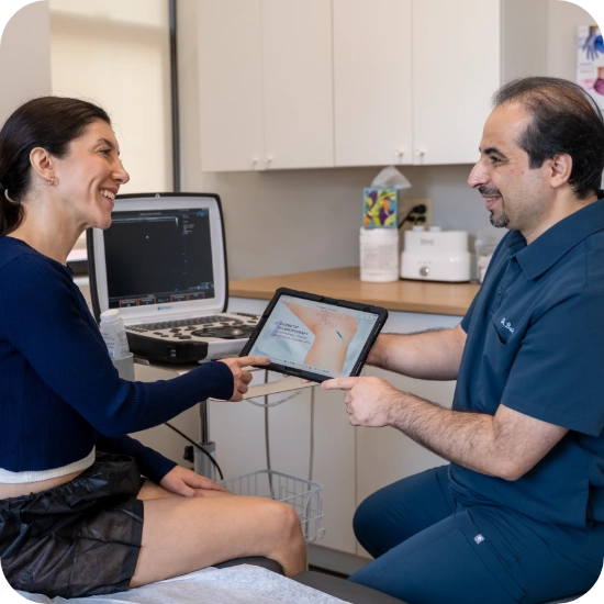 Double certified Dr Kamran Saraf Dr. Mohsen Basiri consults with a female patient in a clinical office setting, utilizing a tablet device to review diagnostic imaging or educational content related to venous conditions. The provider demonstrates and explains clinical findings while the patient sits attentively. The background displays a medical office equipped with an ultrasound machine, diagnostic software on a laptop, and various medical supplies.